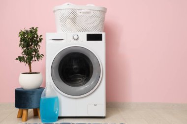 Pembe duvarın yanında sepetli, çiçekli ve deterjanlı modern çamaşır makinesi.