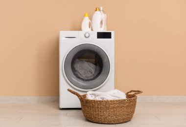 Bej duvarın yanında çamaşır, sepet ve deterjan olan modern çamaşır makinesi.
