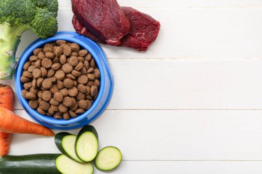 Sebzelerin yanında sağlıklı hayvan mamasıyla kase ve beyaz ahşap arka planda et.