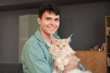 Evde sevimli Maine Coon kedisi olan yakışıklı, genç ve mutlu bir adam.