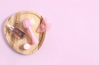 Ahşap tepsi, yüz masajı aletleri, kavanoz ve pembe arka planda bir şişe kozmetik ürünü.