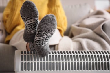 Genç bir kadın, oturma odasında modern konteyner ısıtıcısında bacaklarını ısıtıyor.