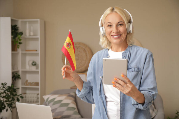 Mature woman in headphones with flag of Spain and tablet computer learning language online at home