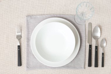 Masada beyaz masa örtüsü, tabak, cam ve çatal bıçak takımı olan minimalist bir ortam.