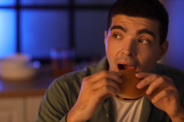 Geceleri mutfakta hamburger yiyen aç genç adam, yakın plan.