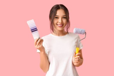 Pembe arka planda boya silindiri ve palet örnekleri olan güzel genç bir kadın.