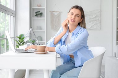 Dizüstü bilgisayarı ve bir fincan sıcak kahveyle evde diş ağrısı çeken genç bir kadın.