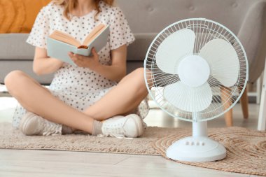 Elinde elektrikli yelpaze olan genç bir kadın oturma odasında oturmuş kitap okuyor.