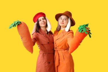 Tavşan kulaklı ve şık paltolu iki genç kadın sarı arka planda oyuncak havuç tutuyor. Paskalya kutlaması