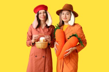 Şık paltolu ve tavşan kulaklı iki genç kadın ellerinde oyuncak havuç ve sepetle sarı arka planda Paskalya ekmeği tutuyorlar. Paskalya kutlaması