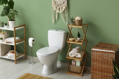 Tuvalet klozeti, raf ünitesi, kağıt tutacağı ve tuvaletteki yeşil duvarın yanında çamaşır sepeti.