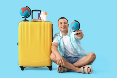 Küresel ve bavullu erkek turist mavi arka planda oturuyor.