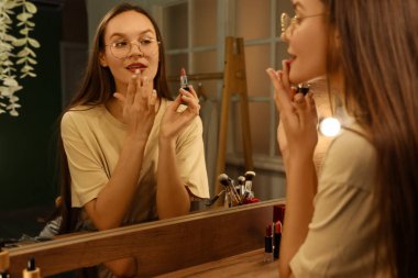 Genç bir kadın gece makyaj odasında aynanın yanına ruj sürüyor.