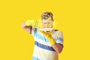 Süngerli genç adam sarı arka planda bulaşık yıkıyor.