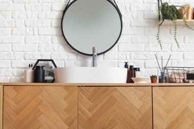 Beyaz tuğla duvarın yanında lavabo kasesi ve banyo aksesuarları olan tahta bir dolap.