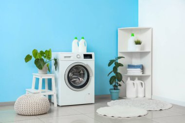 Çamaşır makinesi, raf ünitesi, ev bitkileri ve mavi duvarın yanındaki puf.