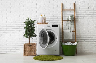 Beyaz tuğla duvarın yanında merdiveni, sepeti ve bitkisi olan modern çamaşır makinesi