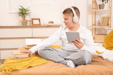 Kulaklık, tablet bilgisayar ve kitaplarla evde oturan küçük bir çocuk.