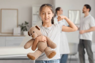 Çocuk doktorunun ofisinde oyuncak ayıyla kaslarını gösteren tatlı bir kız.