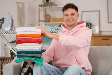 Evdeki kanepede oturan bir yığın temiz elbisesi olan yakışıklı genç adam.