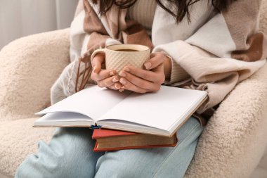 Kitapları, battaniyesi ve kahvesi olan genç bir kadın evdeki koltukta oturuyor.