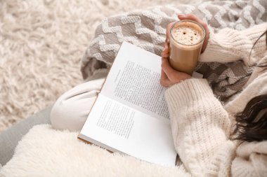 Battaniyeli genç bir kadın, bir fincan kahve ve kitap. Evdeki kanepede oturuyor, yakın plan.
