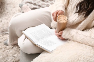 Elinde bir fincan kahve ve kitapla evdeki kanepede oturan genç bir kadın.