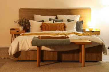 Şık bir yatak odası, ahşap bir bank, rahat bir yatak ve yatağın üzerinde parlayan bir lamba.