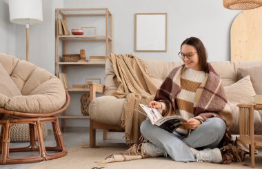 Oturma odasındaki halıda sıcak ekoseli dergi okuyan genç bir kadın.