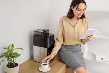 Ofiste kahve yaparken cep telefonu kullanan güzel genç bir iş kadını.