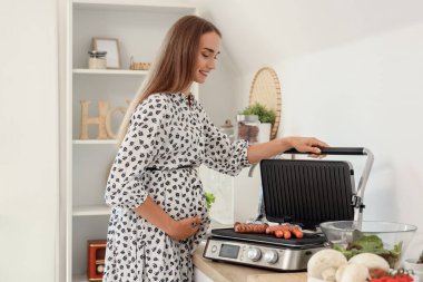 Güzel genç hamile kadın mutfaktaki modern elektrikli ızgarada sosis pişiriyor.