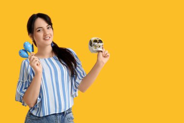 Sarı arka planda boyalı kafatası ve marakas olan güzel genç bir kadın. Meksika Ölüler Günü