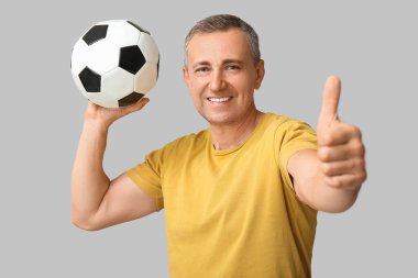 Futbol topuyla yakışıklı, olgun bir adam gri arka planda baş parmak hareketi yapıyor.