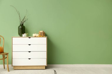 Yeşil duvarın yanında beyaz çekmeceler, balkabakları, mumlar ve ağaç dalları