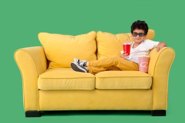 3 boyutlu bardaklı küçük çocuk, yeşil arka planda kanepede film izliyor.