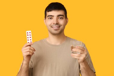 Young man with blister of vitamin A pills and water on yellow background clipart