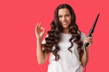 Beautiful young woman with curling iron showing OK on red background