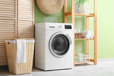 Sepetli modern çamaşır makinesi, raf ünitesi ve yeşil duvarın yanındaki giyinme ekranı. Ev çamaşır odasının içi.