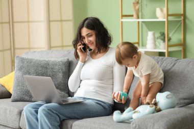 Evde cep telefonuyla konuşurken dizüstü bilgisayarla çalışan tatlı bir bebek ve genç anne.