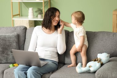 Evde cep telefonuyla konuşurken dizüstü bilgisayarla çalışan tatlı bir bebek ve genç anne.