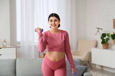 Güzel hamile kadın oturma odasında dambıllarıyla antrenman yapıyor.