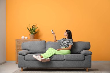 Evdeki turuncu duvarın yanındaki kanepede oturan klimalı uzaktan kumandalı genç bir kadın.