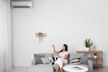 Oturma odasındaki beyaz tuğla duvarın yanındaki koltukta klimalı uzaktan kumandalı genç bir kadın.