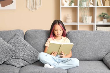 Küçük tatlı Asyalı kız kitap okuyor ve oturma odasında kanepede oturuyor.