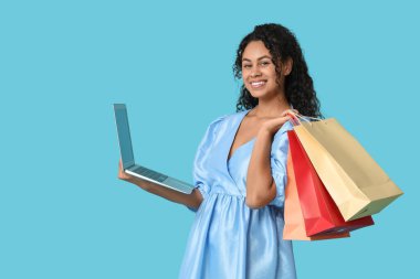 Alışveriş torbaları ve mavi arka planda dizüstü bilgisayarı olan mutlu bir Afro-Amerikalı kadın.