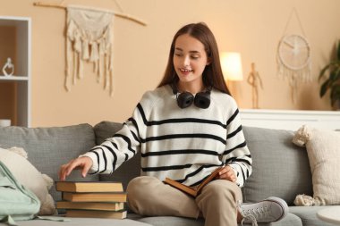 Evdeki kanepede oturan kitap ve kulaklıklarla güzel, mutlu bir bayan öğrenci.