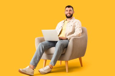 Dizüstü bilgisayarı olan yakışıklı genç adam sarı arka planda koltukta oturuyor.