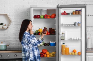 Mutfakta taze yiyeceklerle dolu açık buzdolabının yanında reçel dolu bir kavanozla düşünceli Asyalı kadın.