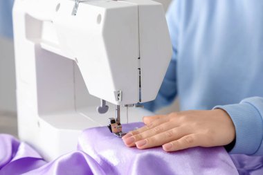 Genç bayan terzi atölyede, yakın plan masadaki leylak kumaş için dikiş makinesi kullanıyor.