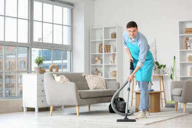 Önlüklü genç adam evde süpürge halısı temizliyor.